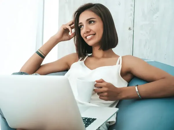 Portrait of beautiful smiling woman dressed in white pajamas. Carefree model sitting on soft bag chair and using laptop.